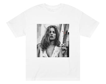 Puede incluir: Una camiseta gráfica en blanco y negro con una mujer de pelo largo, con un top blanco y sosteniendo un cigarrillo. El fondo es una imagen borrosa de una valla de madera.