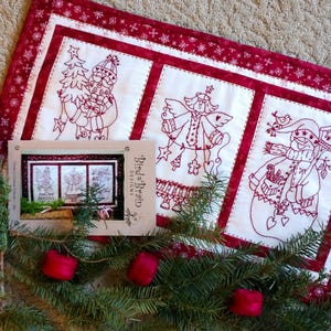 May include: A rectangular fabric wall hanging featuring three white panels with red embroidered holiday figures including a Santa, an angel, and a snowman, framed by a red border with white snowflake patterns, accompanied by a Bird Brain Designs card.