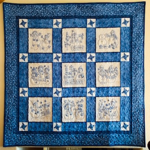 Puede incluir: Una colcha cuadrada con nueve bloques de tela blanca con escenas de muñecos de nieve bordadas en azul, separadas por bandas de tela azul con remolinos y pequeñas piezas de esquina en forma de estrella. Bordeada por una franja azul oscuro.