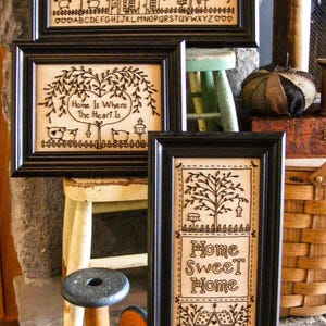 May include: A collection of framed, embroidered art pieces with black frames. The artwork features homes, trees, and the phrases "Bless Our Home" and "Home Sweet Home." The style is rustic and country-themed, with a neutral color palette.