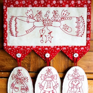 May include: A red and white Christmas wall hanging with three embroidered angels. The top angel is holding a star and two hearts. The bottom three angels are holding hearts and stars.