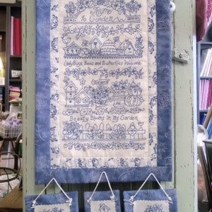 May include: A blue and white fabric wall hanging with the text "Home to Garden" and three smaller matching square hangings. The main piece features embroidered garden scenes and the words "Ladybugs, Bees and Butterflies Welcome".