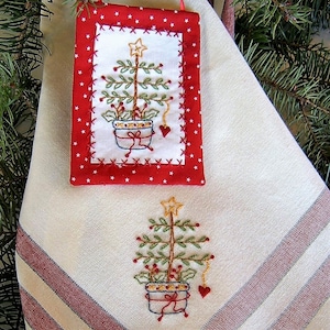 May include: A red fabric ornament with white polka dots and a white embroidered Christmas tree in a pot. The ornament is hanging on a white kitchen towel with red and white stripes and a smaller embroidered Christmas tree in a pot.