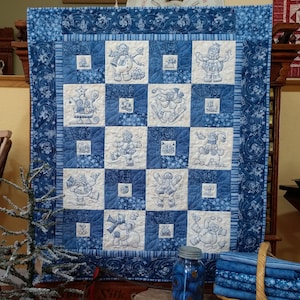 May include: A blue and white quilted wall hanging with a snowflake pattern. The quilt is divided into 16 squares, each featuring a different embroidered snowman design. The quilt is framed with a blue border.