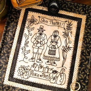 May include: A black and white embroidered Thanksgiving-themed wall hanging with a cream background. The embroidery features two figures, a pumpkin, apples, and the text "Give Thanks!" and "Peace * Plenty".