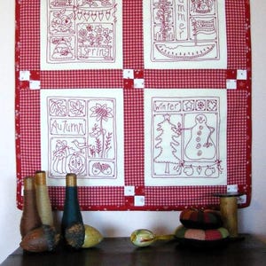 Könnte beinhalten: Ein rot-weiß karierter Quilt mit vier bestickten Tafeln, die die Jahreszeiten darstellen: Frühling, Sommer, Herbst und Winter. Der Quilt wird über einem dunklen Holzschrank mit dekorativen Gegenständen ausgestellt.