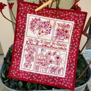 May include: A square, quilted wall hanging with a red floral pattern border. The center features four embroidered panels with flowers and the text "Take Time To Smell The Flowers!" in red thread. A wooden clothespin holds it in place.