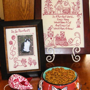 Può includere: Un ricamo incorniciato con il testo "In A Purrfect World... Every Home Would Have A Cat And Every Cat Would Have A Home!" è esposto accanto a una foto incorniciata di un gatto. Sono visibili anche un giocattolo a forma di topo floreale rosso e bianco e una ciotola per cibo per gatti.