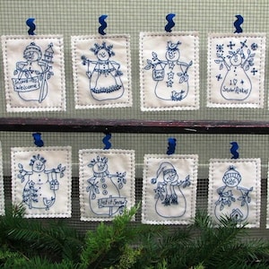 May include: Eight white fabric ornaments with blue embroidery of snowmen with different designs. The ornaments are hanging on a wire mesh background. The designs include "Snowbirds Welcome", "Bird Seed", "Let it Snow", and "I Love Snowflakes".