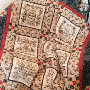 May include: A beige and black quilt with a patchwork design featuring embroidered houses and floral motifs. The quilt is made up of squares with different designs, including a house with the word "Home" above it, a house with the alphabet above it, a house with the words "Blessings of Home" above it, and a house with the words "Welcome Friends & Family" above it.