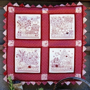 May include: A quilted wall hanging with four embroidered panels depicting the seasons. Each panel features floral and seasonal elements, with text labels for Spring, Summer, Autumn, and Winter. The quilt has a red and white color scheme.