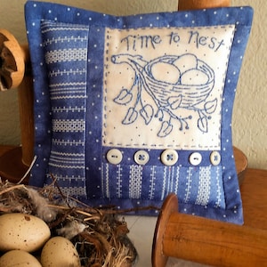 May include: A blue and white pillow with a white embroidered design of a bird's nest with eggs and the text "Time to Nest". The pillow has a row of white buttons along the bottom.