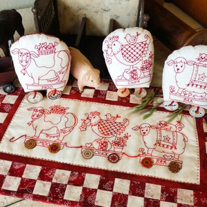 May include: A set of five embroidered fabric toys depicting farm animals on wheels. The toys are a cow, a chicken, a sheep, and two more cows. The toys are arranged in a line on a red and white checked fabric. The toys are all white with red embroidery. The toys have the words "Moo", "Cluck", and "Baa" embroidered on them.