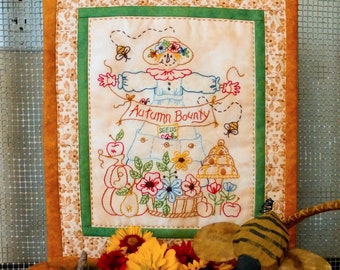 May include: A hand-stitched embroidered wall hanging featuring a scarecrow with the text "Autumn Bounty" and a basket of fall harvest items. The background is a cream-colored fabric with a brown and green border.