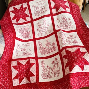 May include: A red and white quilt with a star pattern and embroidered images of cats. The quilt features various cat-themed designs, including cats playing, cats in a garden, and cats with hearts. The text "Love Me Love My Cat!" is embroidered on the quilt.