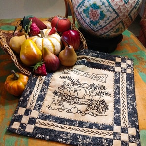 Puede incluir: Un camino de mesa acolchado de color crema con un patrón floral en blanco y negro. El camino de mesa presenta un diseño bordado en blanco y negro de una cesta llena de fruta y el texto "Simple Blessings".