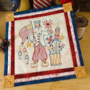 May include: A patriotic-themed embroidered wall hanging featuring a cartoon Uncle Sam holding a flag and a firework. The background is a cream-colored fabric with red, white, and blue borders.