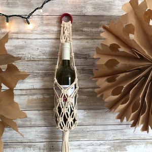 以下が含まれることがあります： クリーム色のロープと赤い木製リングが付いたマクラメワインボトルホルダー。ホルダーは白い木製の壁に掛けられています。