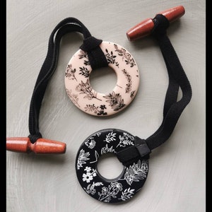 May include: Two decorative hair tie holders. One is black with white floral designs, the other is a light pink with black floral designs. Each has a black elastic band and a wooden toggle.