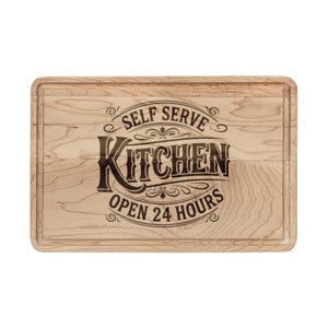 May include: A rectangular wooden chopping board with a juice groove. It features the words "SELF SERVE KITCHEN OPEN 24 HOURS" in black script. The board has a light wood grain pattern and a beveled edge.