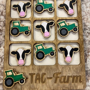 May include: A wooden game board with a green tractor and a black and white cow design. The board has nine squares and says "TAC-Farm" at the bottom.