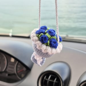 May include: A crocheted car charm featuring a bouquet of blue roses with green leaves, set in a light blue and white holder. The charm hangs from a white cord, adding a touch of handmade charm to any vehicle's interior.