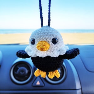 Può includere: Un ornamento a forma di uccello all'uncinetto fatto a mano, con testa bianca, corpo nero e becco e zampe gialle. L'uccello ha grandi occhi neri ed è appeso a un filo nero. Lo sfondo mostra un cruscotto di un'auto e una spiaggia.