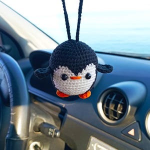 Op de afbeelding: Een gehaakte pinguïn autohanger met zwart en wit bont, oranje voeten en een witte snavel. De pinguïn hangt aan de achteruitkijkspiegel van een auto.