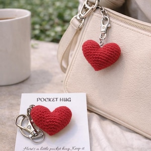 May include: A red crocheted heart keychain hanging from a cream-colored purse. A second heart keychain rests on a card that reads "POCKET HUG". The card includes a message about keeping the hug close. A white mug is in the background.