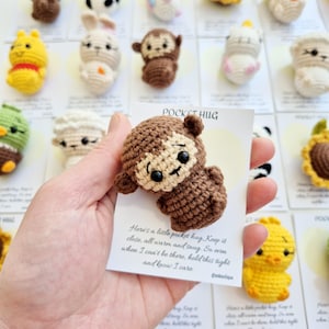 May include: A crocheted brown monkey with a cream-colored face and black eyes is held in front of a card that says "Pocket Hug". Other crocheted animals are in the background, including a yellow chick and a white bunny.