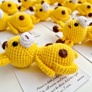 May include: Crocheted yellow giraffe toys with brown spots and white accents. The handmade stuffed animals are lying on white cards. The text on the card reads "Here's a little pocket..." and "Elsmboutique".