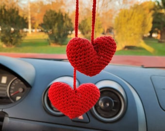 May include: Two red crocheted hearts hanging from a rearview mirror in a car. The hearts are made of yarn and have a textured surface.