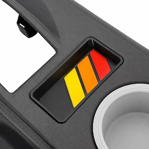 Op de afbeelding: Een zwart middenconsole-paneel voor auto's met een getextureerd oppervlak, een ronde witte bekerhouder en een rechthoekige uitsparing met drie diagonale strepen in de kleuren geel, oranje en rood.