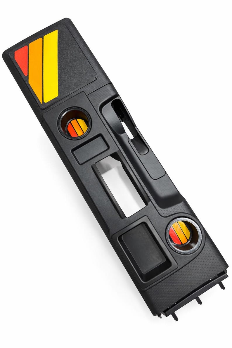 Pu&ograve; includere: Console automobilistica nera con portabicchieri e vani portaoggetti integrati. Presenta un emblema decorativo con strisce rosse, arancioni e gialle. Include uno slot per la leva del cambio.