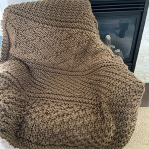 May include: A brown knitted blanket with a textured pattern is draped over a chair. The blanket has a variety of stitch patterns, creating a cozy and inviting look. The neutral color complements the room's decor.