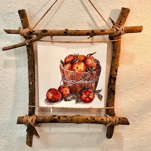 Puede incluir: Una pintura de acuarela de un cubo de madera lleno de manzanas rojas, con tres manzanas que se caen sobre un fondo blanco. La pintura está enmarcada con ramas naturales y cuerda.