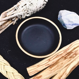 May include: A black ceramic bowl with a gold rim, surrounded by white sage, palo santo sticks, and a blue crystal. The items are arranged on a black surface.