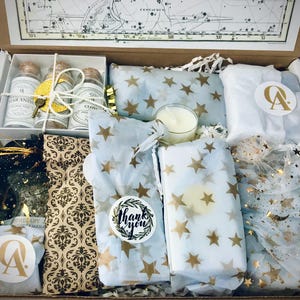 May include: A gift box filled with various items, including small glass bottles, a candle, and tissue paper with a star pattern. The box is lined with brown paper and has a white label with the text "Thank you".