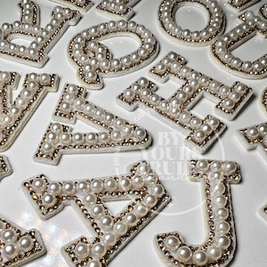 May include: A set of pearl and gold letter patches. The letters are in uppercase and are surrounded by a circle of pearls. The letters are on a white background.