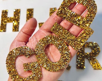 Può includere: Toppe di lettere dorate realizzate con strass scintillanti. Le lettere compongono la parola "GOLD".