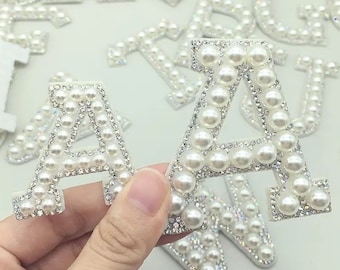 Puede incluir: Un conjunto de parches de letras con adornos de perlas y diamantes de imitación en forma de las letras A y W. Los parches son blancos y tienen un acabado brillante.