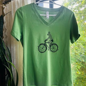 May include: A light green V-neck t-shirt featuring a black graphic print of a creature riding a bicycle. The shirt has short sleeves and a standard hem, with a white brand label visible inside the neckline on the back of the garment.