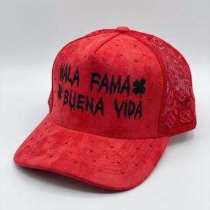 May include: A bright red baseball cap featuring a suede-like texture with scattered red rhinestones. The front panel displays the black embroidered text MALA FAMA BUENA VIDA with a clover symbol. The back panels consist of red mesh fabric.