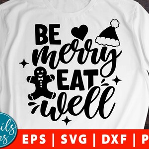 Pode incluir: Camiseta branca com texto preto que diz "Be merry eat well". O texto está em uma fonte festiva. Há um gráfico de homem de gengibre e um gráfico de chapéu de Papai Noel.