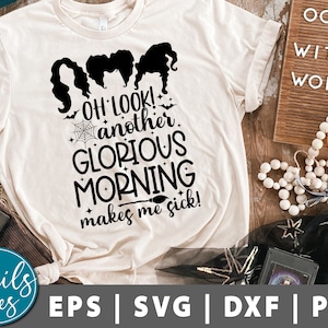 May include: A white t-shirt with a black graphic design featuring a cartoon image of three witches with the text "OH LOOK! another GLORIOUS MORNING makes me sick!" The text "OOH, WITCHY WOMAN" is visible on a wooden sign in the background.