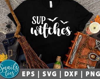 May include: Black t-shirt with white text that says "Sup Witches" and three white bats. The shirt is on a table with a broom, a book, and other Halloween decorations.