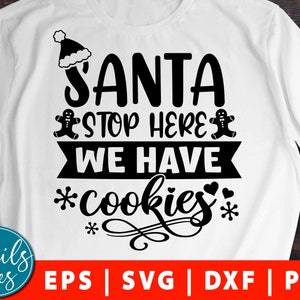 May include: A white t-shirt with black text that reads "Santa Stop Here We Have *cookies*" with a gingerbread man on either side of the text. The text is in a festive font and includes a snowflake and hearts.