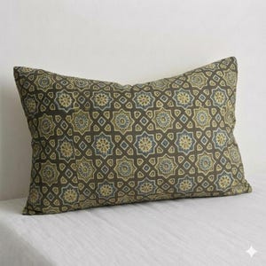 Può includere: Cuscino decorativo rettangolare con un motivo geometrico ripetuto in verde oliva, blu e giallo su sfondo marrone scuro. Il cuscino è posto su una superficie bianca.