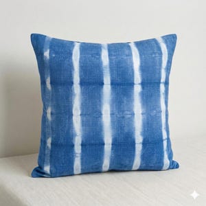 May include: Square blue pillow with a white striped pattern. The pillow has a solid blue border and a textured appearance. The pillow is a decorative home accent.