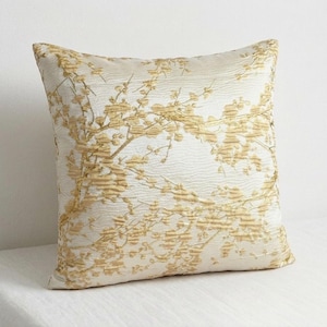 May include: Square decorative pillow with a cream-colored background and a gold floral pattern. The pillow has a textured surface and a neutral color palette, suitable for various home decor styles.